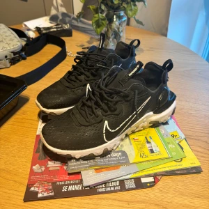 Nike React Vision 43 - Svarta Nike react vision sneakers med vit sula och vit swoosh. Ovandelen är i syntetmaterial med perforerade detaljer för extra andningsförmåga. Skorna har snörning, rund tå och en chunky, dämpad sula med unika mönster. Perfekta för dig som gillar streetstyle. Som nya jätte bra skick