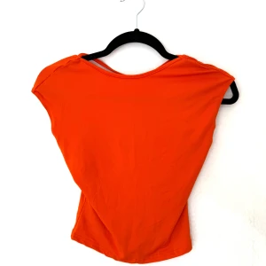 Orange topp med öppen rygg - Snygg orange topp i storlek XS. Modellen är ärmlös med öppen rygg. Tunt och stretchigt material som sitter tajt och framhäver figuren. Lite nopprig