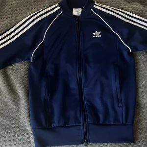 Adidas blå tröja med vita ränder - Säljer en klassisk marinblå Adidas tröja i storlek S. Tröjan  har vita ränder längs ärmarna, dragkedja framtill och två fickor. Tillverkad i polyester med Primeblue-material. Perfekt för en sportig och avslappnad stil. 