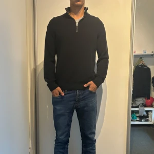 Svart stickad tröja med half zip - Svart stickad tröja med halv dragkedja i halsen. Tröjan har lång ärm och en klassisk, stilren look som passar perfekt till jeans. Materialet är mjukt och ger en skön känsla, perfekt för kyligare dagar.