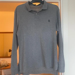 Grå långärmad pikétröja Polo Ralph Lauren - Snygg grå långärmad pikétröja från Polo Ralph Lauren med klassisk krage och två knappar framtill. Tröjan har broderad logga på bröstet och är gjord i mjuk bomull. Perfekt för en clean och stilren look.