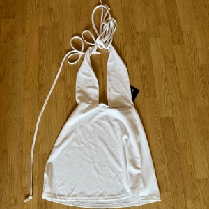 Vit halterneckklänning PrettyLittleThing - Supersnygg vit kort klänning från PrettyLittleThing med halterneck och djup v-ringning. Klänningen har knytband i nacken och är ärmlös med en lätt utsvängd kjol. Perfekt för sommarens alla tillfällen!