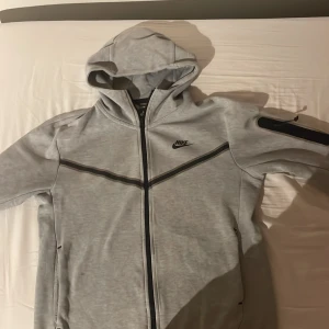 Grå Nike hoodiejacka med dragkedja - Snygg grå hoodiejacka från Nike med hel dragkedja och svarta detaljer. Jackan har huva, svarta linjer över bröstet och en praktisk ficka med dragkedja på ärmen. Perfekt för en sportig och avslappnad stil.