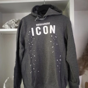 Grå hoodie Dsquared2 ICON XXL - Snygg grå hoodie från Dsquared2 i storlek XXL med vit 'ICON'-text och coola vita stänkdetaljer framtill. Klassisk huva med snörning och mjukt material som känns skönt mot huden. Perfekt för dig som gillar streetwear och vill sticka ut.