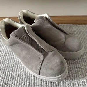 Grå mocka slip-on sneakers från Eytys - Stilrena slip-on sneakers från Eytys i ljusgrå mocka med chunky vit platt sula. Skorna har elastisk panel framtill och en lila detalj baktill. Insidan är fodrad i vitt och sulan har tydlig struktur undertill. Perfekta för dig som gillar minimalistisk och modern stil.