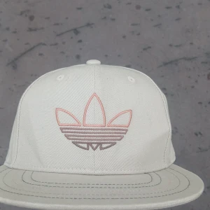 Vit keps från Adidas Originals - Snygg vit keps från Adidas Originals med broderad Trefoil-logga i ljusrosa och beige toner framtill. Klassisk modell med platt skärm och justerbar passform. Tillverkad i bomull för en bekväm känsla och stilren look.