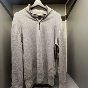 Grå half zip tröja från BOSS - Snygg grå stickad tröja från BOSS med half zip och hög krage. Tröjan har lång ärm och en diskret struktur i materialet. Perfekt för dig som gillar stilrena och klassiska plagg med en modern touch.