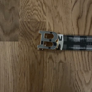 Burbbery bälte - Snyggt bälte med rutigt mönster i svart och grått. Bältet har ett stort, guldigt B-format spänne med räfflad yta. Tillverkat i skinn med en stilren och modern look, perfekt för att lyfta din outfit med en statement-detalj.