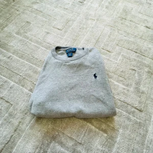 Grå tröja från Polo Ralph Lauren - Klassisk grå tröja från Polo Ralph Lauren (Storlek 10-12år) i mjuk pima-bomull. Tröjan har rund halsringning, diskret broderad logga på bröstet och en clean look som funkar till allt. Perfekt för dig som gillar stilren och enkel design.