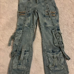 Dolls kill cargo pants - Säljer ett par ljusblå baggy cargopants i jeansmaterial med flera stora fickor, dragkedjor och långa remmar med nitar längs benen. Byxorna har en riktigt streetig vibe och är perfekta för dig som gillar detaljer och oversized look.