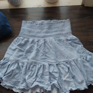 Ljusblå volangkjol från 157, S - Supersöt ljusblå kortkjol från 157 med smock i midjan och volang nertill. Kjolen har små blommiga detaljer i rosa och vitt, vilket ger en romantisk vibe. Perfekt för dig som gillar en feminin och lekfull stil.