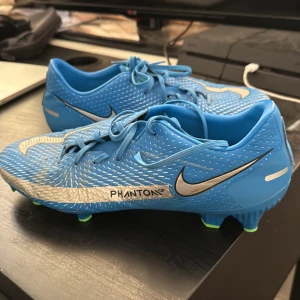 Nike Phantom GT2 blå fotbollsskor - Nike Phantom GT2 fotbollsskor i blått med silverfärgade detaljer och vita mönster. Skorna har snörning, rund tå och gröna dobbar för bästa grepp på planen. Tillverkade i syntetmaterial för lätt känsla och bra bollkontroll. Perfekta för dig som vill sticka ut på matchen.