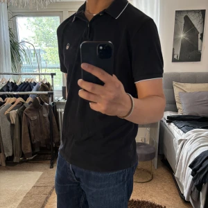 Svart pikétröja Sail Racing L - Svart pikétröja från Sail Racing med vit logga på bröstet och tunna vita detaljer på krage och ärmslut. Klassisk pikékrage med två knappar. Skön passform och tillverkad i bomull, perfekt för en clean och stilren look.