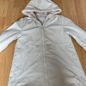 Beige regnjacka med fuskpäls från Zara storlek 140/ 10 år - Snygg beige regnjacka från Zara med mjukt fuskpälsfoder i luvan och insidan. Jackan har dragkedja framtill, två fickor med dragkedja och huva. Size 10 barn 140cm