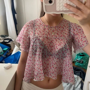 Blommig transparent blus i rosa och vit - Supersöt blommig blus i transparent material med korta ärmar och croppad passform. Toppens vita bas har små rosa och röda blommor över hela tyget. Luftig och flowy modell som ger en romantisk vibe, perfekt för varma dagar.