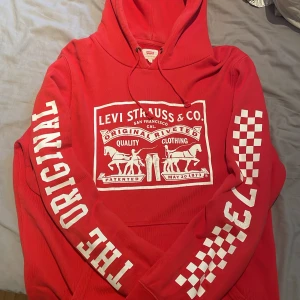 Röd hoodie från Levi's med tryck - Röd hoodie från Levi's med stort vitt tryck på bröstet och texten THE ORIGINAL på ena ärmen. Snygga rutiga detaljer på ärmarna och klassisk huva med snörning. Perfekt för dig som gillar streetstyle och vill sticka ut lite extra.