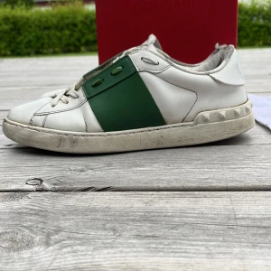 Valentino Garavani sneakers med gröna detaljer - Säljer ett par snygga Valentino Garavani sneakers i vitt med breda gröna detaljer och diskret logga på plösen. Skorna har en klassisk rund tå och platt sula, perfekt för dig som gillar exklusiva märken och stilren design.