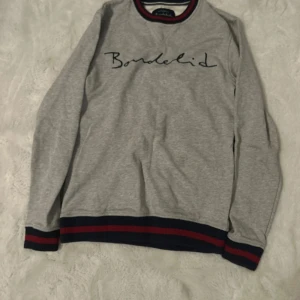 Grå tröja från Bondelid med ränder - Snygg grå långärmad tröja från Bondelid med marinblå och röda ribbade detaljer vid hals, ärmslut och nederkant. Tröjan har ett stilrent tryck med Bondelid-loggan framtill och är gjord i mjuk bomull. Perfekt för en chill och sportig look.