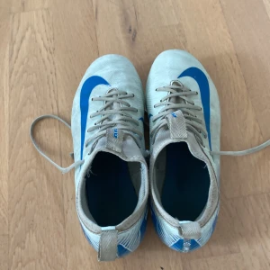 Nike Mercurial vita och blå fotbollsskor vapor med bra skick  - Säljer ett par Nike Mercurial fotbollsskor i vitt med blå detaljer och snörning. Skorna har platt sula med dobbar för bra grepp på planen och är tillverkade i syntetmaterial. Perfekta för dig som vill sticka ut på fotbollsplanen!