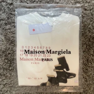 Maison Margiela T-shirt - Vit tröja från Maison Margiela Paris med tryck av siffror och svarta boots framtill. Snygg och stilren design, perfekt för dig som gillar exklusiva streetwear-plagg. Tröjan har rund hals och är i mjuk bomull. 