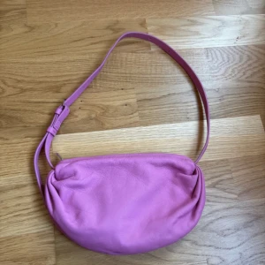 Rosa axelväska i skinn från COS - Säljer en snygg rosa axelväska från COS i mjukt skinn. Väskan har en enkel, rundad form och justerbart axelband. Stängs med dragkedja och har ett foder i bomull. Perfekt för dig som vill ha en färgklick till din outfit.