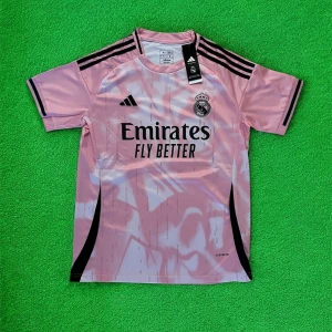 Rosa Real Madrid fotbollströja Adidas - Snygg rosa Real Madrid fotbollströja från Adidas med svart logga och klubbmärke på bröstet. Tröjan har svarta detaljer på axlar och ärmar, samt trycket 'Emirates Fly Better' på framsidan. Tillverkad i lätt och ventilerande material.