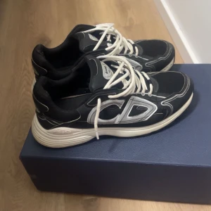 Dior b30 - Snygga svarta Dior sneakers med vita snören och kontrasterande vita detaljer. Skorna har en chunky sula och stora CD-loggor på sidan. Ovandelen är i en mix av skinn och textil för en modern look. Perfekta för dig som vill sticka ut med exklusiva sneakers. Box och skosnören kommer med 