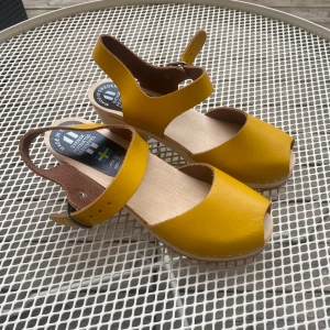 Gula svenska trätoffel-sandaler - Snygga sandaler i klargult skinn med träbotten och öppen tå. Justerbart spänne i metall och klassisk svensk design. Perfekta för dig som vill sticka ut med färg och gillar retrovibbar. Passar till både jeans och klänning under varma dagar. I princip helt oanvända, liten liten defekt på ena hälen, pris kan diskuteras!