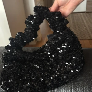 Svart glittrig handväska med paljetter - Säljer en svart handväska täckt med glittriga paljetter. Väskan har en rundad form och ett vadderat handtag, samt dragkedja upptill. Perfekt för dig som vill sticka ut med en cool accessoar. Rymmer mobil, plånbok och småsaker.