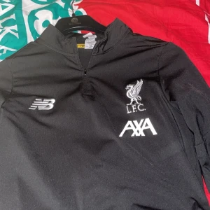 Liverpool långärmad träningströja New Balance - Snygg svart långärmad träningströja från Liverpool FC med broderad L.F.C.-logga och AXA-sponsor på bröstet. Tillverkad i lätt och funktionellt material, perfekt för träning eller när du vill representera ditt lag. Dragkedja vid halsen och klassisk passform. Köpt i Liverpool i shoppen