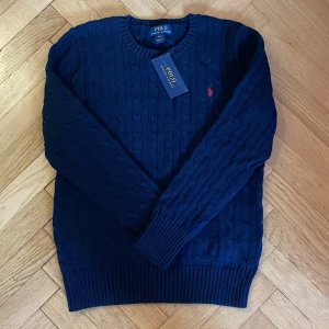 Stickad Polo Ralph Lauren tröja - En klassisk mörkblå stickad Polo Ralph Lauren tröja. Köpt för många år sedan men är aldrig använd; inga defekter. Tröjan är i barnstorlek och passar dig som är mellan 14-16 år eller 150-161cm lång.