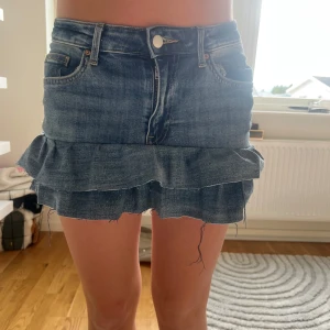 Jeanskjol med volang - Supersöt kort jeanskjol med volangkant och råa, fransiga avslut. Klassisk blå denim med fickor fram och bak samt bälteshällor. Perfekt för dig som gillar en trendig och avslappnad stil.
