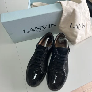 Svarta sneakers från Lanvin i mocka och lack - Snygga svarta sneakers från Lanvin med ovandel i mocka och glansig lackad tå. Klassisk rund tå, svarta skosnören och diskret branding på sulan. Skorna levereras med originalkartong och dustbag. Perfekta för dig som gillar stilrena och lyxiga detaljer.