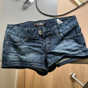 Mörkblå jeansshorts från Guess - Snygga mörkblå jeansshorts från Guess med klassisk femficksdesign och coola nitar på bakfickan i form av Guess-loggan. Shortsen har låg midja och är gjorda i stretchigt denim för en bekväm passform. 