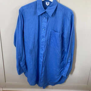 Blå linneskjorta från H&M, strl 34 - Snygg blå skjorta i linne från H&M i storlek 34. Oversized.  Klassisk krage, långärmad med knappar framtill och en bröstficka. Perfekt oversized look och skönt material som andas. Enkel och stilren design som passar till många outfits. Fläck på ena framsidan, se bild.