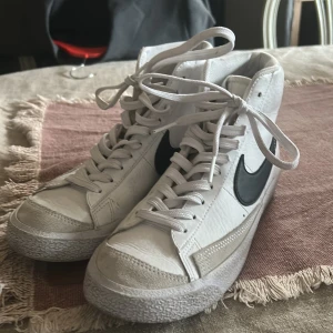 Nike Blazer Mid vita sneakers - Snygga vita Nike Blazer Mid sneakers med svart swoosh och klassisk retrostil. Skorna har högt skaft, snörning och är gjorda i skinn med detaljer i mocka framtill. Perfekta för dig som gillar streetwear och vill ha en tidlös look.