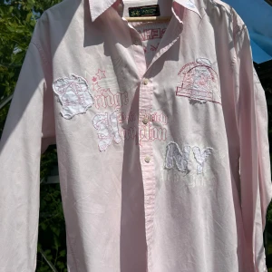 Rosa skjorta La Martina Bridgehampton L - Snygg rosa skjorta från La Martina med broderade och tryckta detaljer på bröstet och ryggen. Stora loggor, text och emblem i vitt och rosa ger en sportig och unik look. Klassisk krage, knäppning framtill och lång ärm. Perfekt för dig som gillar statement-plagg.