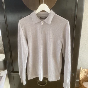 Ljusgrå ziptröja från J.Lindeberg - Snygg ljusgrå tröja från J.Lindeberg med hel dragkedja framtill och klassisk krage. Tröjan är stickad i mjukt material och har långa ärmar samt ribbade muddar. Perfekt för dig som gillar stilren och enkel design. Ett pittelitet hål under högra armhålan. Nypris: 1300