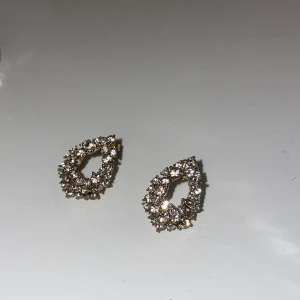 Glittriga droppformade örhängen - Säljer ett par snygga örhängen i droppform med glittrande stenar. Basen är i guldton och hela örhänget är täckt av små, gnistrande kristaller som ger en lyxig känsla. Perfekta för att lyfta vilken outfit som helst med extra bling. 