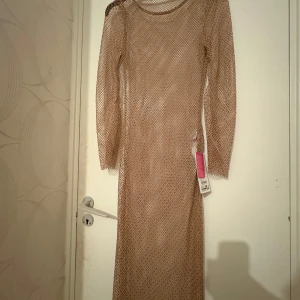 Beige nätklänning med lång ärm - Trendig beige nätklänning med lång ärm och rund halsringning. Klänningen är figurnära och har ett transparent nätmaterial som ger en cool och edgy look. Perfekt att styla över andra plagg för en unik outfit.