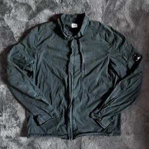 C.P. Company overshirt jacka - Säljer en mörkgrön overshirt från C.P. Company med klassisk krage och knappstängning. Perfekt till lite kyliga sommarkvällar. Storlek L, skick 8/10 inga defekter men solblekt, pris går att diskutera✅