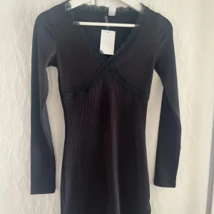 Svart ribbad klänning med spetsdetaljer - Svart långärmad klänning med ribbad struktur och v-ringning. Spetsdetaljer vid halsringningen ger en extra touch. Klänningen har en kroppsnära passform och är perfekt för dig som gillar stilrena plagg med fina detaljer.