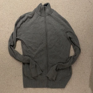 Grå stickad full zip tröja - Säljer en stilren grå ziptröja med hög ribbad krage och hel dragkedja framtill. Tröjan har långa ärmar och ribbade muddar vid ärmslut och nederkant. Perfekt som lager under jackan eller till vardags.