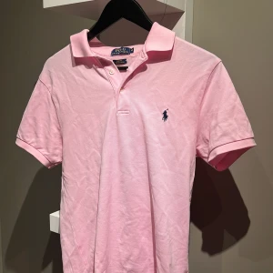 Rosa pikétröja från Polo Ralph Lauren - Säljer en klassisk rosa pikétröja från Polo Ralph Lauren i slim fit. Tröjan har korta ärmar, krage och knappar framtill samt den ikoniska broderade loggan på bröstet. Tillverkad i stretchig bomullsmix för extra komfort. Köpt på NK för 1400, säljer pga för liten.