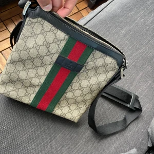 Gucci axelväska  - Säljer en Gucci axelväska med klassisk GG-monogram och grönt/rött band på framsidan. Väskan har svarta detaljer, justerbar axelrem och dragkedja. Perfekt för dig som vill ha en snygg accessoar nu på sommaren.