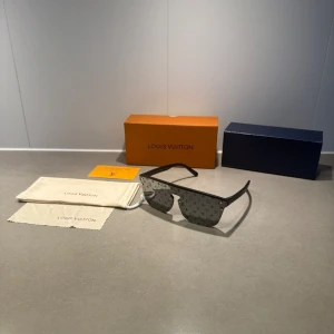 Svarta LV glasögon  - Stilrena svarta solglasögon från Louis Vuitton med LV-monogram på glasen. Bågarna är i svart plast och har en modern, kantig form. Perfekt accessoar för dig som vill sticka ut med exklusiv design.