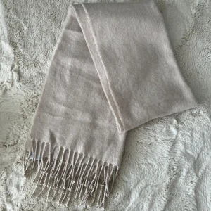 Beige halsduk från H&M - Säljer en beige halsduk från H&M med klassiska fransar i ändarna. Halsduken är mjuk och passar perfekt för att hålla värmen under kyliga dagar. Enkel att matcha med olika outfits.