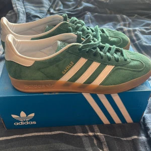 Adidas Gazelle Indoor gröna sneakers - Säljer ett par gröna Adidas Gazelle Indoor sneakers med vita ränder och detaljer. Skorna har klassisk snörning, gummisula och är tillverkade i mocka med vit hälkappa. Perfekta för dig som gillar retrostil och vill sticka ut med färg.