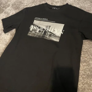 Svart t-shirt med Brooklyn Bridge-tryck från Denim Day - Säljer en svart t-shirt från Denim Day med stort tryck av Brooklyn Bridge i vitt och grått på bröstet. Klassisk passform och rund halsringning. Perfekt för dig som gillar streetstyle och grafiska motiv.