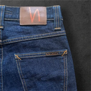 Mörkblå jeansbyxor från Nudie Jeans - Säljer ett par mörkblå jeansbyxor från Nudie Jeans med klassisk femficksdesign och kontrastsömmar. Byxorna har raka ben och hög midja, tillverkade i slitstarkt denim. Perfekta för dig som gillar tidlös stil och bra passform.